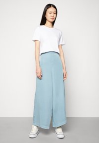 Calças largas de cor azul clara feitas de tecido suave, com uma racha frontal e um brilho subtil, combinadas com uma t-shirt branca simples de manga curta.