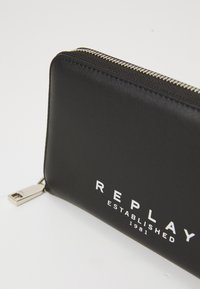 Černá kožená peněženka se stříbrným zipem, na přední straně s bílým logem "REPLAY ESTABLISHED 1981" a kovovým prvkem.
