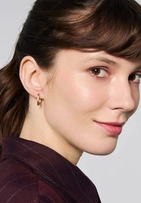 Złoty kolczyk w kształcie koła noszony na lewym uchu, charakteryzujący się gładką, refleksyjną powierzchnią i okrągłym wzorem. Ułożony na tle ciemnofioletowej tkaniny.