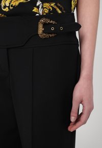 Μαύρο παντελόνι ψηλής μέσης με χρυσή πολύπλοκη αγκράφα. Ο ύφασμα έχει απαλή υφή, με κομψές πλευρικές πτυχώσεις και εφαρμοστή σιλουέτα.