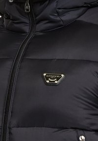Nærbillede af sort polstret jakke med frontlynlås og trekantsformet metal-logo patch med teksten "Alessandro Zavetti Milano."