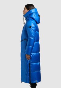 Ilgas mėlynas pūkinis paltas su aukštu apykakliu, užtrauktukais užsegamomis kišenėmis ir ryškiu siuvimu. Medžiaga blizgi ir izoliuota šilumai.