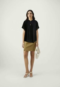 Top preto de mangas curtas com frente plissada e detalhe em forma de chave. Combinado com uma saia mini bege e sandálias de salto beija-flor, segurando uma bolsa branca.