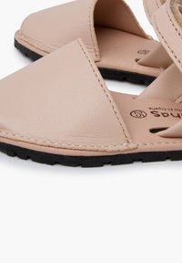 Beige nahkakengät avonaisilla varpailla ja ommelluilla reunoilla. Musta kumipohja, litteä muotoilu ja kaksoishihna-ysityks across the foot.