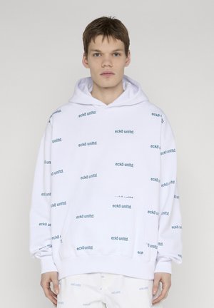 Młody mężczyzna w białej bluzie z kapturem i spodniach z powtarzającym się logo "eckō unltd." na jasnoszarym tle.