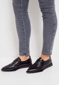 Chaussures en cuir noir avec un bout pointu, un laçage et des coutures décoratives. Portées avec un jean gris à coupe ajustée sur un fond clair.