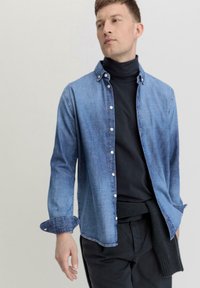 Denimhemd in lichtblauw, met knopen aan de voorkant, lange mouwen met opgerolde manchetten. Draag het over een zwarte coltrui, in combinatie met donkere broeken.