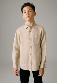 Beige langärmliges Hemd aus leichtem Stoff, mit durchgehendem Knopfleistenverschluss, Kragen und einer Brusttasche, kombiniert mit einer schwarzen Hose.