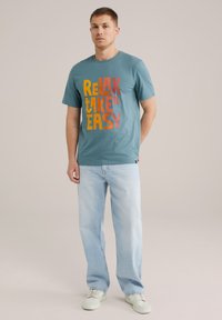 Tengerészkék pamut t-shirt, amelyen merész, színes felirat található: "RELAX TAKE IT EASY." Világoskék, laza szabású farmernadrággal és fehér sportcipővel párosítva.