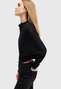 Maglione corto nero in maglia con strisce orizzontali testurizzate. Abbinato a jeans neri a vita alta. Modella posata di profilo.