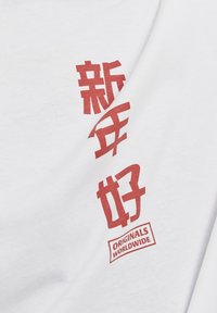 Balta marškinėliai su raudonu tekstu, kuriame yra kinų simboliai ir "ORIGINALS WORLDWIDE" logotipas. Minkšta medvilnės medžiaga, su laisvu kirpimu.