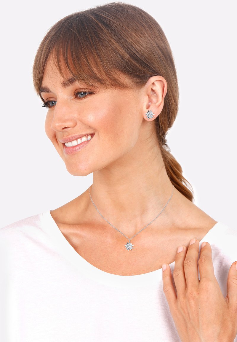 Orecchini a forma di fiocco di neve argentati e collana coordinata, caratterizzati da una delicata catena e un intricato design di pendente a fiocco di neve. Indossati su una maglietta bianca.
