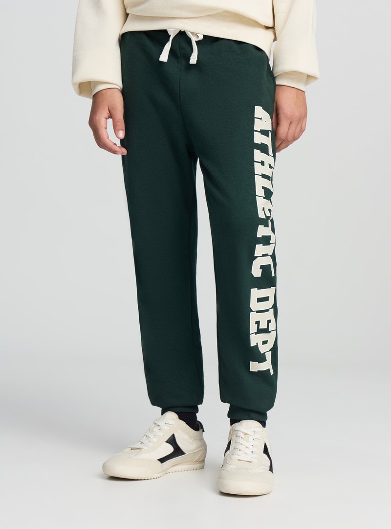 Ciemnozielone spodnie dresowe z białym napisem „ATHLETIC DEPT” wzdłuż lewej nogawki, wyposażone w elastyczny pas i regulowany sznurek.