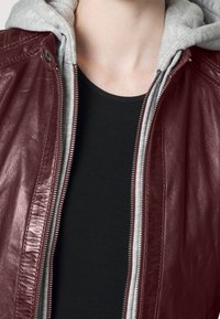 Giacca di pelle bordeaux con fodera grigia con cappuccio, zip argentata e motivo trapuntato sulle spalle, indossata sopra una maglia nera.