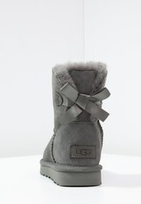 Botte en suède gris avec une doublure en fourrure douce, un ruban décoratif à l'arrière et une semelle texturée avec une marque UGG sur le côté.