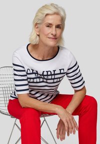 Striped white and navy T-shirt with short sleeves, featuring text design. Paired with red pants. Model seated on a wire chair.