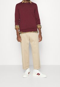 Pull bordeaux porté sur une chemise à carreaux jaune avec des manches retroussées, associé à un pantalon beige raccourci et des baskets blanches à rayures rouges.