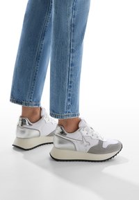 Sneaker bianchi con accenti argentati, punta arrotondata e pannelli grigi testurizzati. Presentano una suola spessa e un design con lacci, abbinati a jeans blu.