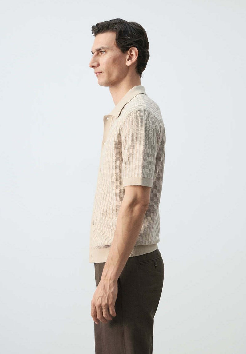 Homme debout en chemise tricotée beige à manches courtes avec col et pantalon marron, regardant vers la gauche sur un fond clair uni.