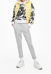 Lehká žlutá péřová vesta přes černobílou mikinu se vzorem zebry, doplněna šedými tepláky a bílými teniskami.