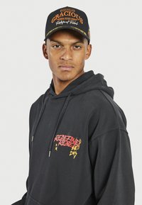 Schwarzer Kapuzenpullover mit rotem und gelbem grafischem Text. Begleitet von einer schwarzen Mütze mit orangefarbenem Stickerei "GRACIOUS" und "Refined Rebel".