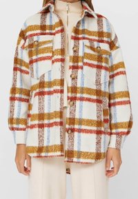 Mujer con una chaqueta de cuadros en blanco, rojo, mostaza, azul y marrón, con botones y bolsillos en el pecho con solapa, sobre una camiseta de cremallera color beige.