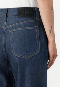 Dark blue denim jeans featuring a straight cut, single back pocket, and a stitched Calvin Klein logo patch above the waistband.