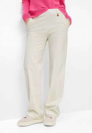 Persoon draagt crèmekleurige wijde broek met zijzakken, beige sneakers en een felroze top met lange mouwen, staand tegen een witte achtergrond.