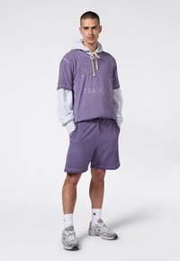 Mężczyzna stojący z rękami w kieszeniach, ubrany w purpurowe szorty i zbyt dużą purpurową bluzę z krótkim rękawem nałożoną na szary longsleeve, w szarych sneakersach.