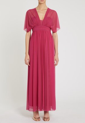 Donna che indossa un abito lungo fino a terra in chiffon rosa scuro con maniche a pipistrello trasparenti, vita raccolta e gonna plissettata, abbinato a tacchi aperti dorati.