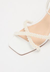 Sandalo con tacco bianco caratterizzato da cinturini incrociati adornati con piccole perle. La suola è beige con una punta quadrata, dal texture liscia.