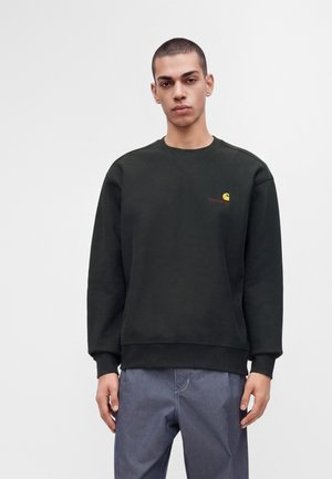 Млад мъж, облечен в червена блуза Carhartt и сини панталони, стои срещу светъл неутрален фон с неутрално изражение.