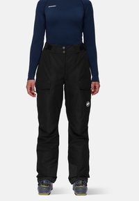 Pantalons de ski noirs imperméables avec poches cargo, taille ajustable et ourlets renforcés. Portés avec un haut à manches longues bleu marine et des bottes de ski grises.