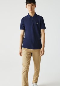 Polo bleu marine avec une finition texturée, patte de boutonnage à trois boutons et un accent logo vert. Associé à un pantalon beige clair et des baskets blanches.