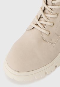 Beige lær ankelsko med rund tå, teksturert overflate, metalliske snørehull og lysfarget robust såle. Lissene er lyse og litt glitrende.