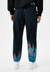 Černé denimové kalhoty s plamennými grafikami v modré a červené na lemech. Mají dvě zadní kapsy. Nosené s bílými teniskami.