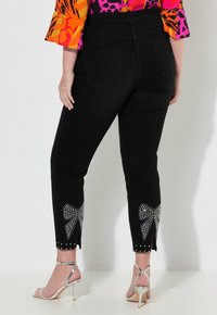 Černé denimové kalhoty s tapered střihem, zdobené třpytivými kamínkovými mašlemi na lemech a stříbrnými detaily. Viditelné zadní kapsy.