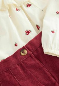 Biała bluzka z czerwonym kwiatowym haftem w zestawieniu z bordowymi spodniami o fakturze. Posiada elastyczne mankiety i dekoracyjny guzik na pasku.