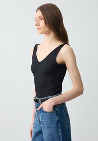Femme aux longs cheveux bruns portant un haut noir sans manches et un jean bleu, une main dans la poche, tournée vers la gauche.