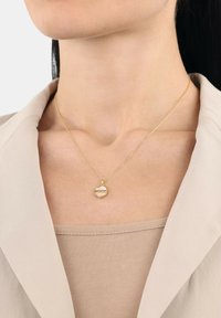 Guldhalsband med en rund berlock med ingraverad text, h ängande på ett tunt kedja, bärs över en beige topp och en ljusfärgad kavaj.