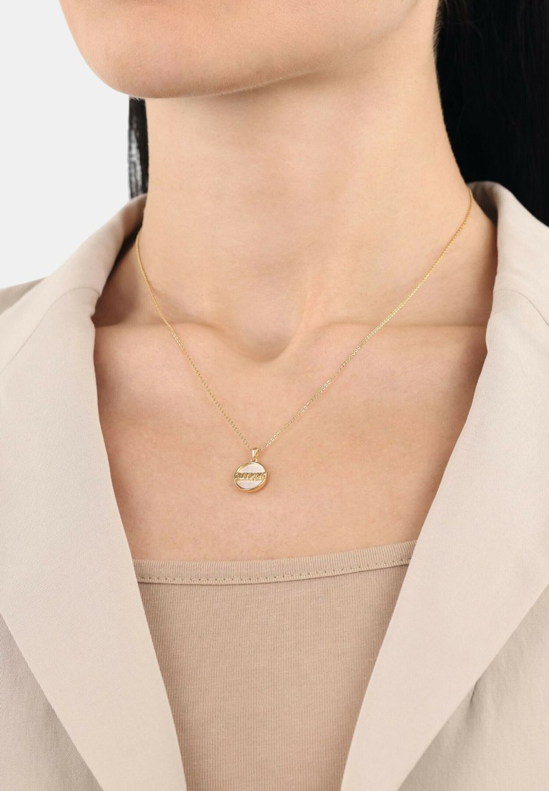 Guldhalsband med en rund berlock med ingraverad text, hängande på ett tunt kedja, bärs över en beige topp och en ljusfärgad kavaj.