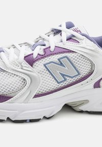 White athletic shoe with purple accents, mesh panels, and textured rubber sole. Features prominent "N" logo on side and white laces.