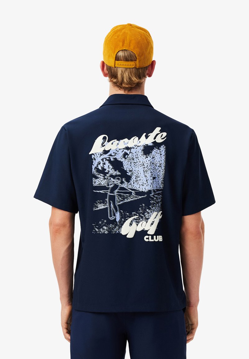 Camicia blu navy a maniche corte con una stampa testurizzata sulla schiena che raffigura un golfista; presenta la scritta bianca "Lacoste Golf Club."