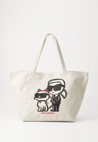 Bežs kanvas iepirkumu soma ar melnu grafiku, kurā attēlots cilvēks saulesbrillēs un kaķis. Somai ir izturīgas rokturi, vienkārša forma un sarkans teksts.