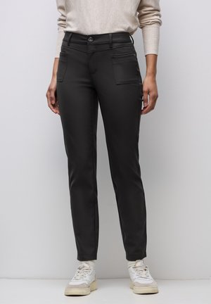 Kobieta ubrana w dopasowane czarne spodnie z przednimi naszywanymi kieszeniami, beżowy sweter, białe skarpetki i białe trampki, stojąca na tle gładkiej białej ściany.