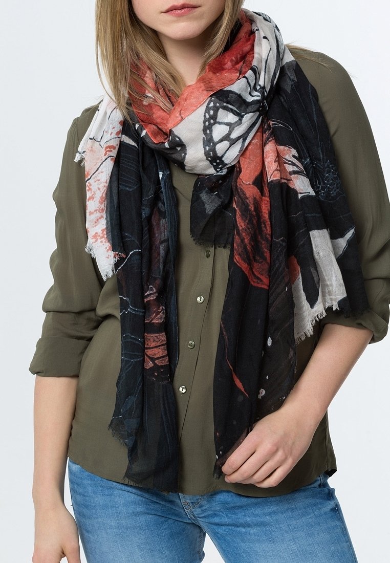Femme portant une chemise kaki boutonnée, un jean bleu et une grande écharpe noire, blanche et rouge à motifs, enroulée autour de son cou.