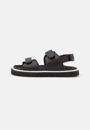 Sandalias negras con dos correas ajustables, una suela plana con costura blanca y una plantilla acolchada para mayor comodidad.