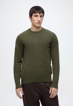 Ung mand iført en olivengrøn kabelstrikket sweater og mørkebrune bukser står mod en ensartet hvid baggrund.
