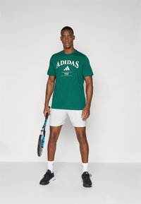 Camiseta de algodão verde com o logótipo "ADIDAS" em creme, combinada com calções brancos e ténis de desporto pretos. O modelo segura uma raquete de ténis.