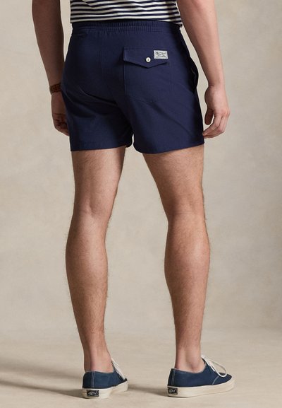 Άνδρας με σκούρες μπλε βερμούδες με πίσω τσέπη και σκούρα μπλε casual παπούτσια, στέκεται σε ουδέτερο μπεζ φόντο.